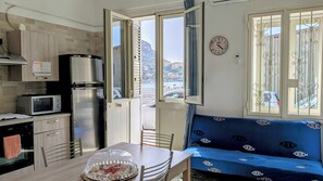 Uma cozinha compacta com micro-ondas, frigorífico e fogão. Há uma mesa de jantar com cadeiras e um sofá azul com vista para o mar.