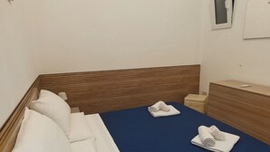 Un llit ben fet amb llençols blancs i un sobrell de color blau, un capçal de fusta, una petita taula de nit amb una làmpada i una finestra amb vistes.
