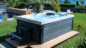 Un jacuzzi con cubierta de madera y escalones, rodeado por una piscina y abundante vegetación.
