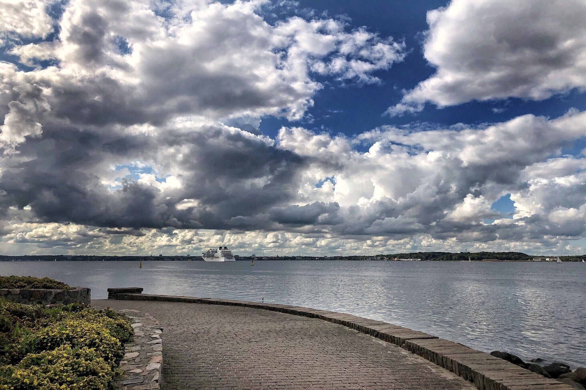 Eine Uferpromenade mit einem gepflasterten Weg, einem großen Gewässer und einem Kreuzfahrtschiff in der Ferne.