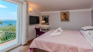 Ein Hotelzimmer mit Bett, Schreibtisch, Stuhl, Fernseher, einem Gemälde und einem Balkon mit Meerblick.