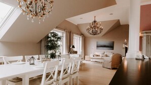 A spacious dining area with a high ceiling and a chandelier, a cozy living room with a sofa and a TV, and a large window providing natural light.