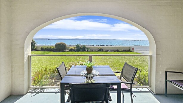 Een eethoek met een tafel en stoelen, een potplant en uitzicht op zee door een rondboogvenster.