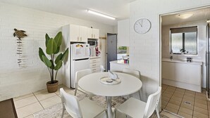 Een compacte keuken met witte kasten, een magnetron en een koelkast. Er is een eettafel met stoelen en een potplant.