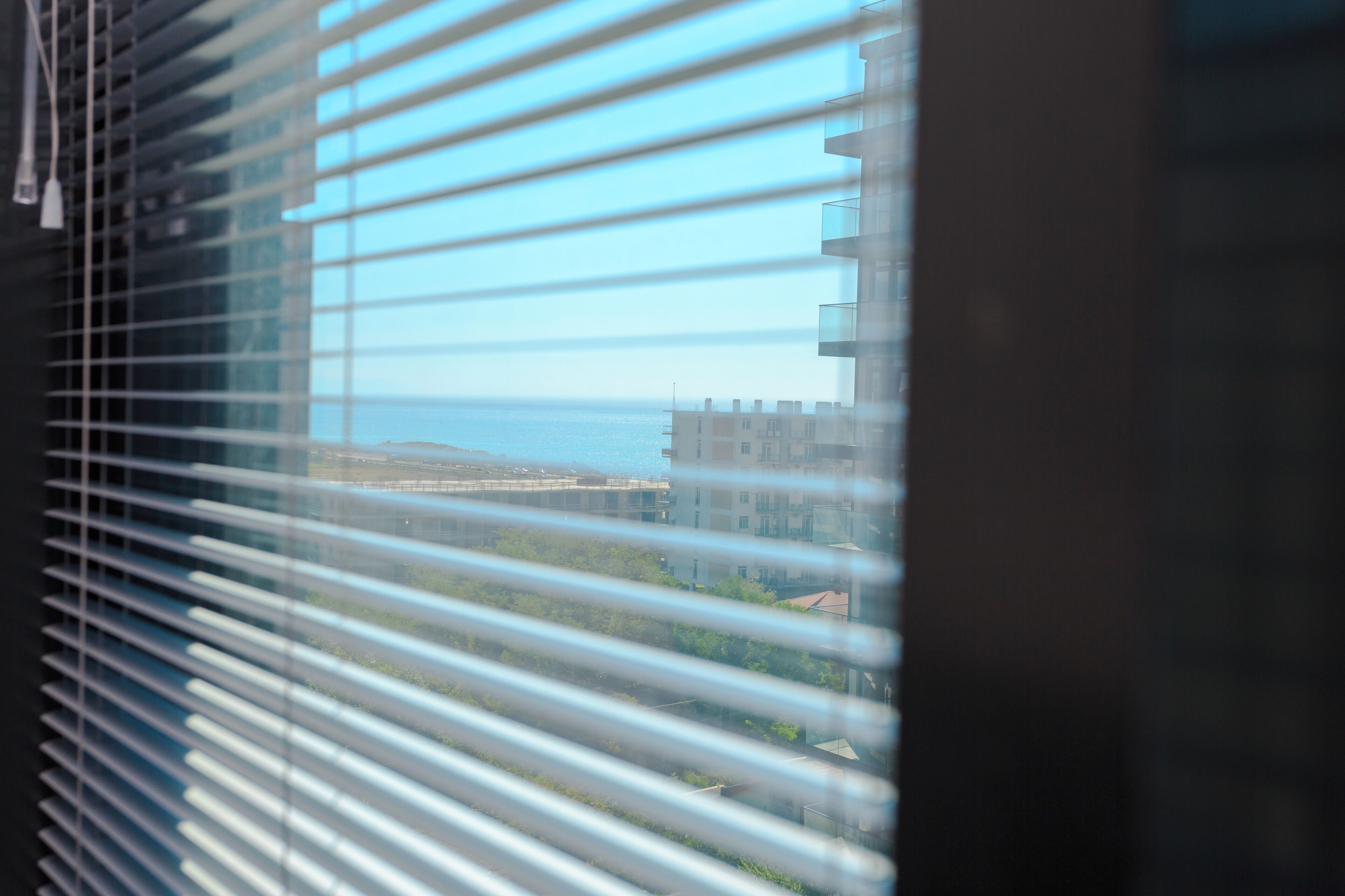 A view through window blinds of a building with a balcony, a clear sky, and a distant body of water.