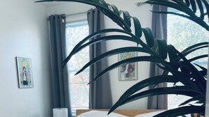 A bed with white bedding, a nightstand with a lamp, a large potted plant, and a window with curtains.