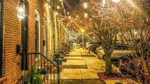 A narrow street lined with brick buildings, illuminated by string lights, with a tree and parked cars visible.