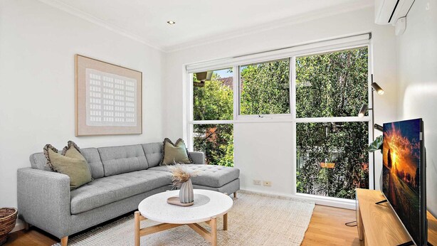 Ein modernes Wohnzimmer mit einer grauen Eckcouch, einem runden Couchtisch und einem großen Fenster mit Blick auf Grünfläche.