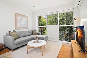 A modern living room with a grey sectional sofa, a round coffee table, and a large window offering a view of greenery outside. - Bright 2-Bed Apartment in South Yarra (South Yarra)