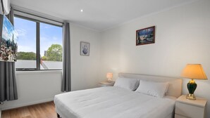 Quarto de hotel com uma cama grande, mesa de cabeceira com abajur, janela com cortinas e um quadro na parede.
