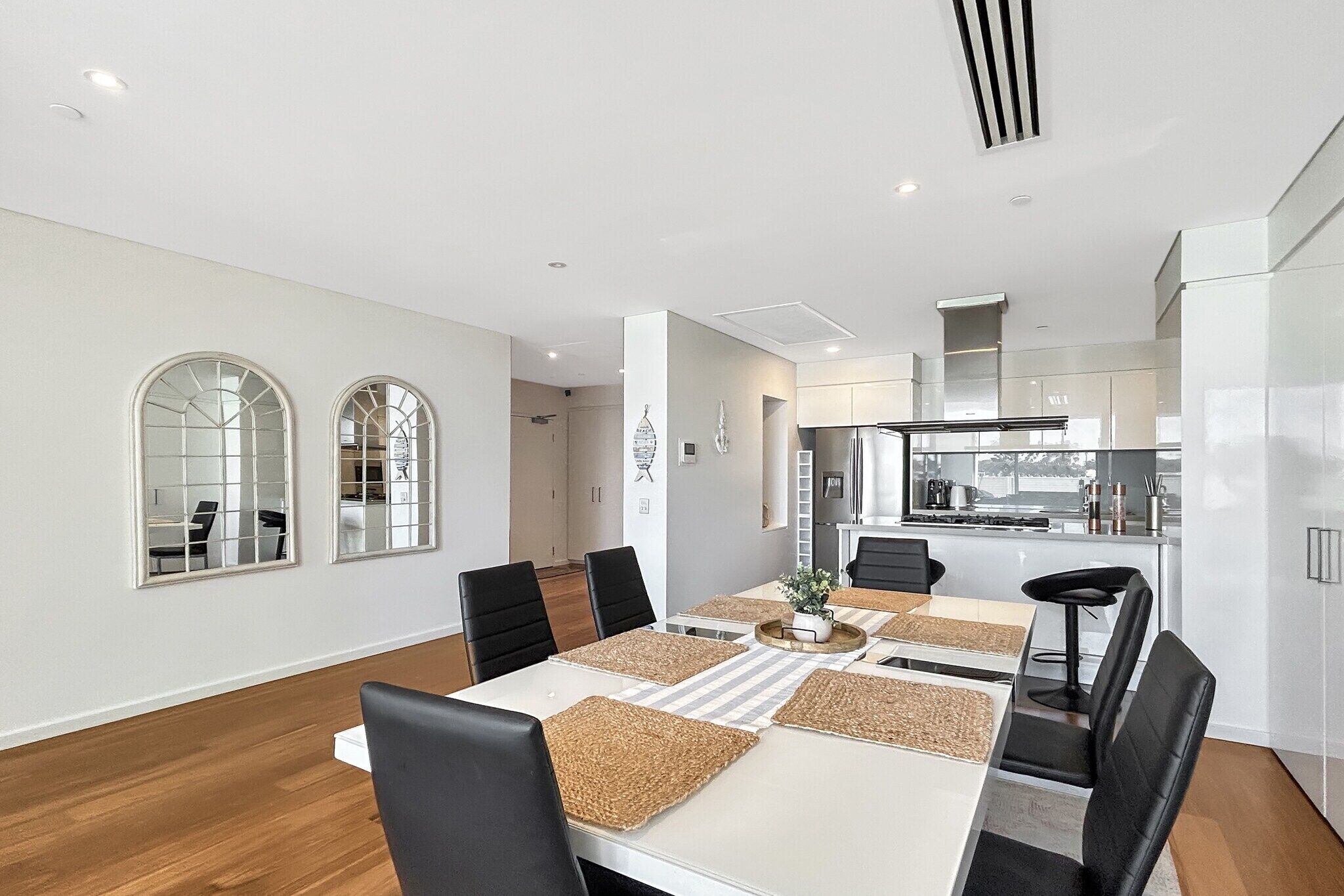 A modern dining area with a white table, black chairs, and a kitchen in the background.