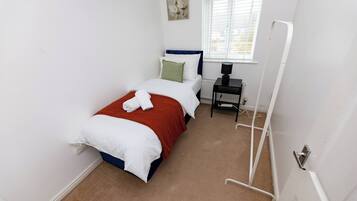 Un dormitorio pequeño y ordenado con una cama individual, un radiador, una ventana con persianas y una puerta con manija.