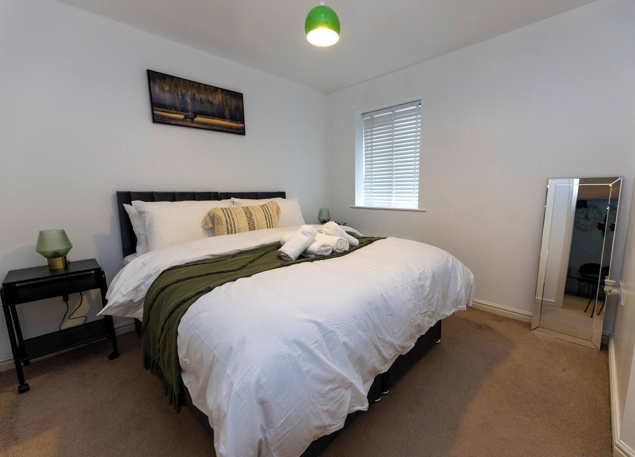 A neatly made bed with white linens, a green throw blanket, and rolled towels. There is a small bedside table with a green lamp, a painting on the wall, and a window with blinds.
