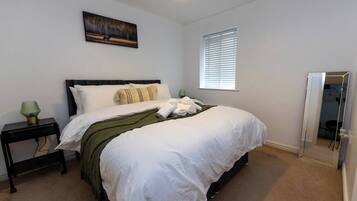A neatly made bed with white linens, a green throw blanket, and rolled towels. There is a small bedside table with a green lamp, a painting on the wall, and a window with blinds.