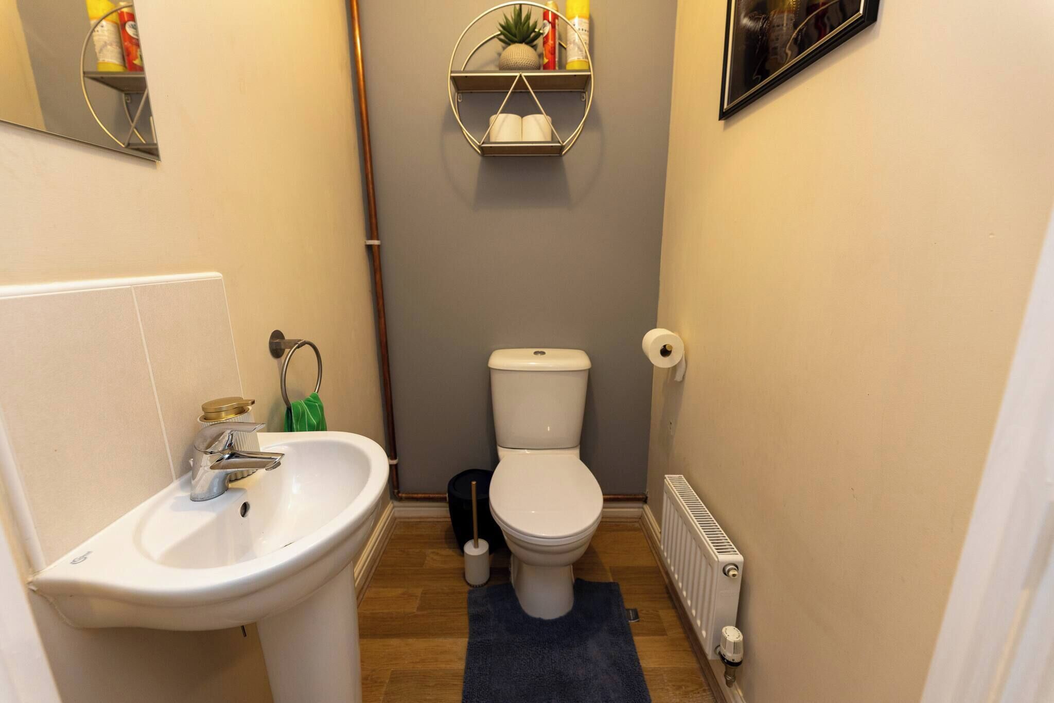 Un baño pequeño con un lavamanos, inodoro y radiador blancos. Hay dos repisas sobre el inodoro con diversos objetos.