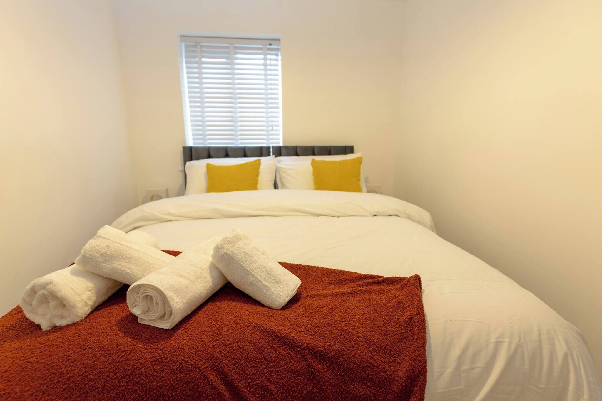 A neatly made bed with white linens and a brown throw blanket, a window with blinds, and a headboard with two pillows.
