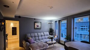 Een moderne woonkamer met een grijze bank, een salontafel en een eettafel. Grote ramen bieden uitzicht op het omliggende landschap.