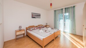 Cama doble con ropa de cama floral, mesita de noche con lámpara, un cuadro en la pared y una ventana con cortinas traslúcidas.