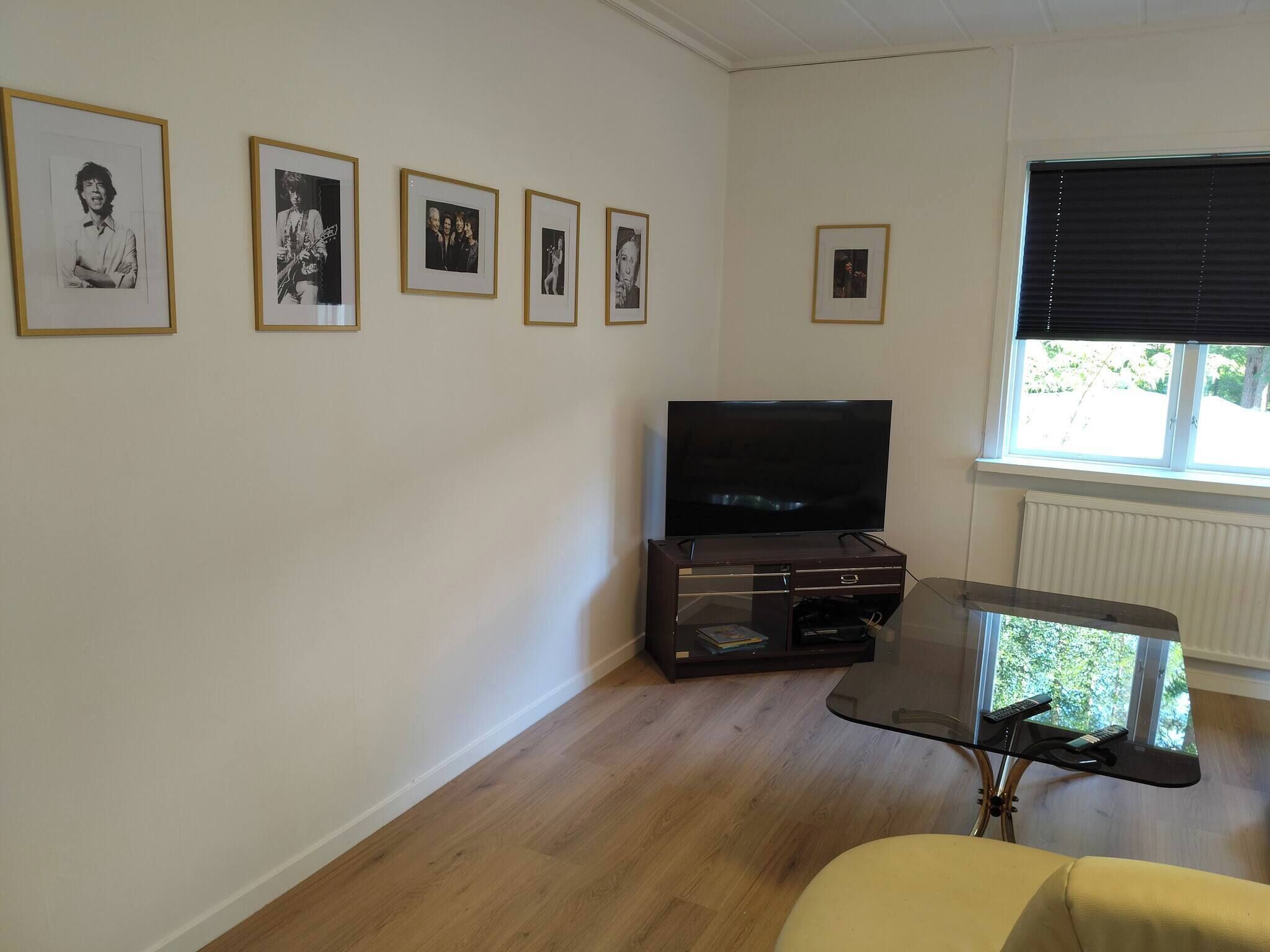 A living room with a flat-screen TV, framed pictures on the wall, a glass coffee table, and a window with blinds.