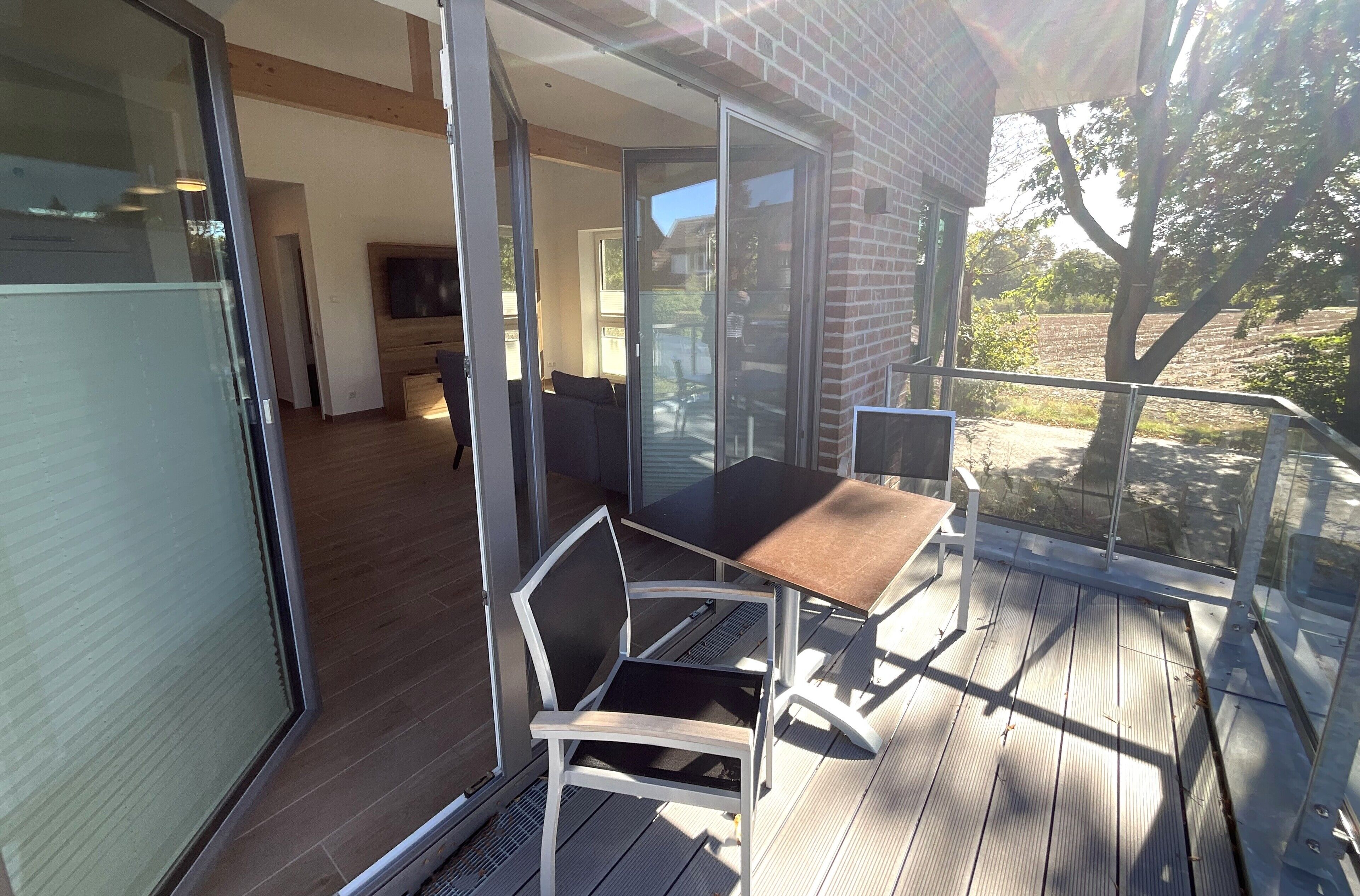A modern outdoor patio with a glass door leading to a living area, a wooden deck, and a view of the surrounding landscape.