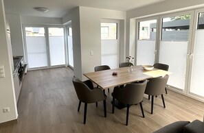 Uma área de refeições moderna com mesa de madeira e seis cadeiras, cozinha ao fundo e grandes portas de vidro deslizantes que dão para um quintal.