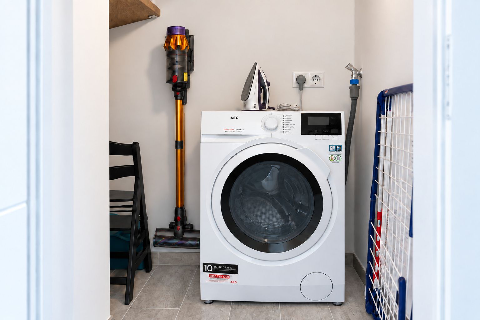 A compact laundry area with a front-loading washing machine, a vacuum cleaner, and a power outlet.