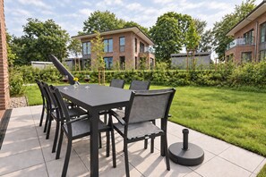 Eine Außenpatio mit einem rechteckigen Tisch und sechs Stühlen, umgeben von einem gepflegten Rasen und einem Backsteinhaus im Hintergrund.