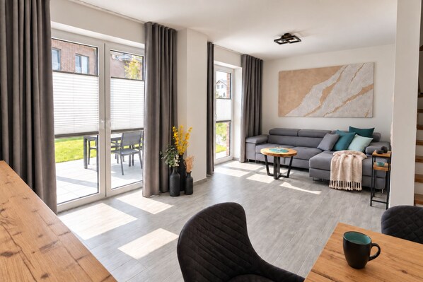 Ein modernes Wohnzimmer mit einem grauen Sofa, einem Holztisch und einem großen Fenster mit Vorhängen.