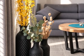 Two black vases with plants on a wooden table, a grey sofa, and a window with sunlight in a living room. - Hello and welcome to the Ackerpulko Vacation Home! (Dangast)