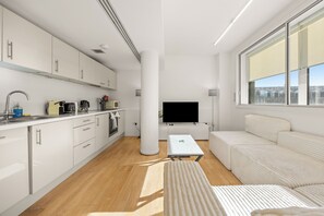 Eine moderne Küche mit weißen Schränken, einem Flachbildfernseher und einer Couch in einem hellen Raum mit großen Fenstern.