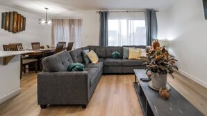 A modern living room with a sectional sofa, a dining area with a wooden table and chairs, and a large window with curtains. - Chiccityescape3bdrm King (Stony Plain)