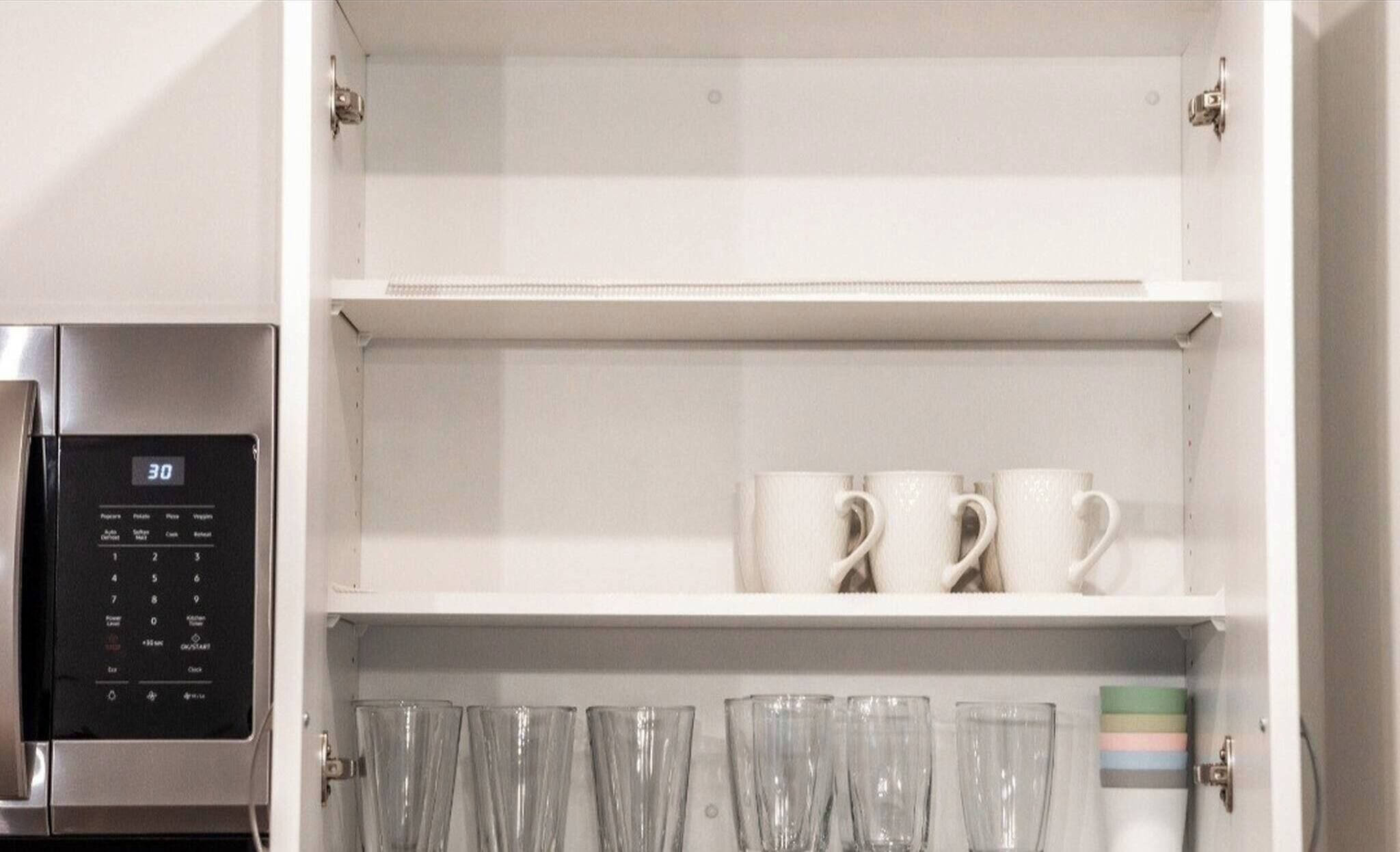A kitchen with a microwave, three white mugs on the top shelf, and several clear drinking glasses on the bottom shelf.