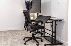 Een moderne werkplek met een zwarte bureaustoel, een bureau met een flatscreenmonitor en een kleine potplant.