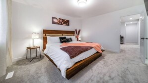 A bedroom with a wooden bed, white bedding, a nightstand with a lamp, and a painting on the wall. - Chiccityescape3bdrm King (Stony Plain)