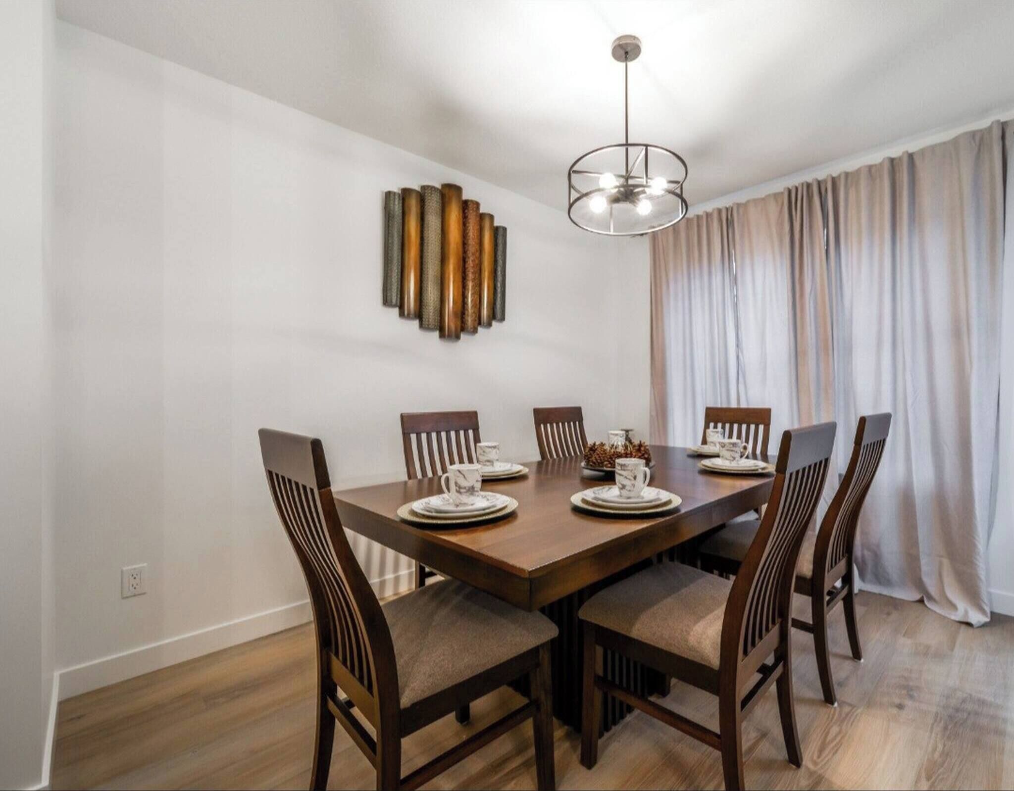 A dining area with a wooden table and chairs, a modern chandelier, and a decorative wall piece.