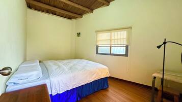 A bedroom with a wooden ceiling, a bed with a blue skirt, a bedside table, and a window with blinds.