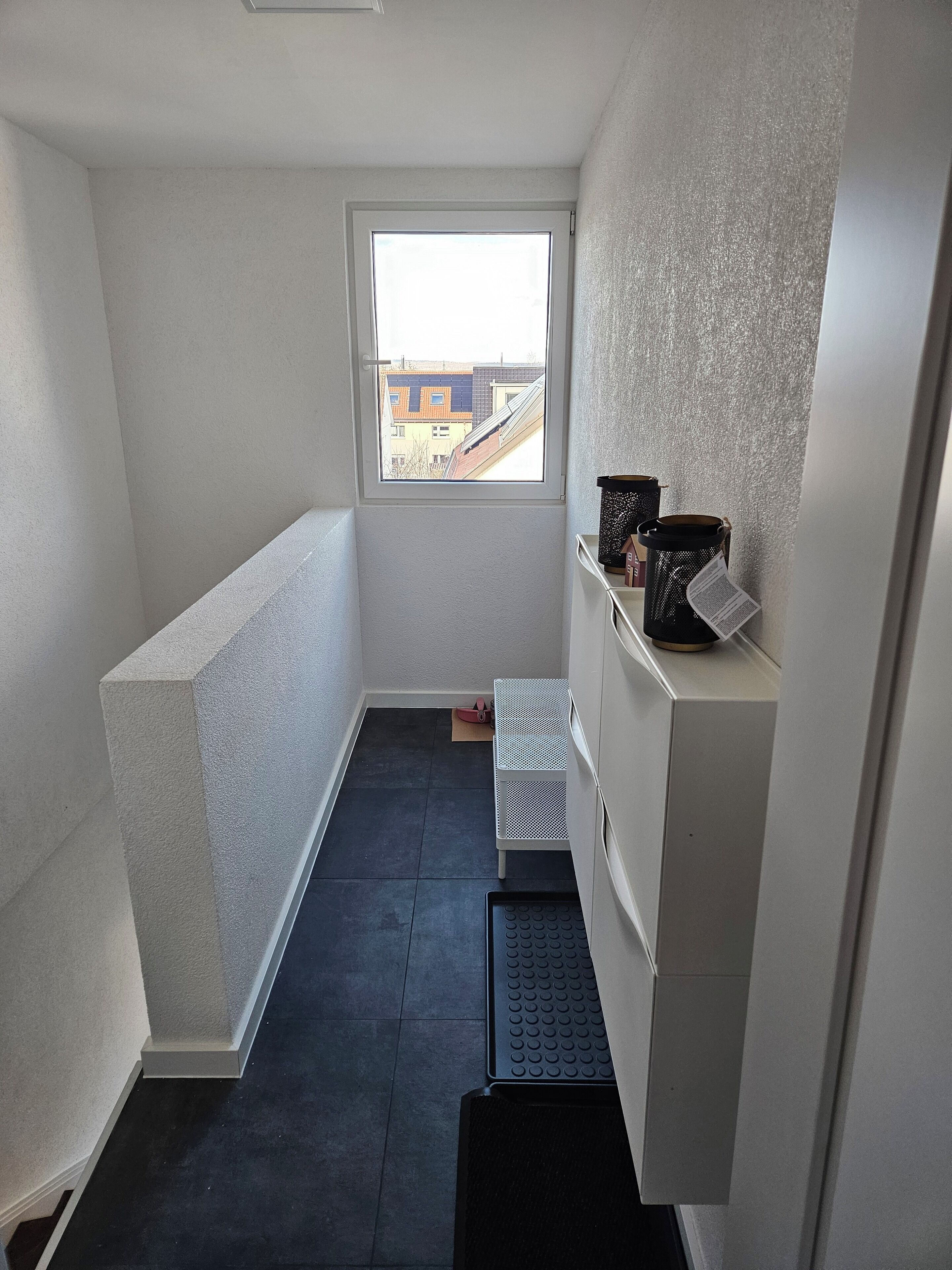 Une pièce étroite et faiblement éclairée, avec une petite fenêtre, une étagère en métal contenant des boîtes de rangement et un tapis noir à l’entrée.