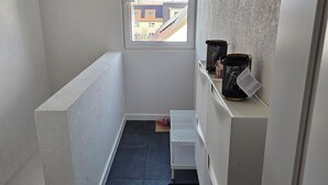 Ein schmaler, schwach beleuchteter Raum mit einem kleinen Fenster, einem Metallregal mit Aufbewahrungsboxen und einer schwarzen Matte am Eingang.