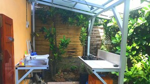 Ein kleiner Outdoor-Kochbereich mit Spüle, Herd und Holzschränken, umgeben von Pflanzen und einer Steinsäule.