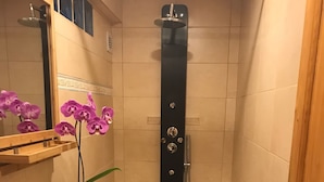 Ein Badezimmer mit einem weißen Waschbecken, einer Holzausstattung, einem Spiegel, violetten Orchideen, einer Dusche mit schwarzem Duschschlauch und einem gelben Teppich.