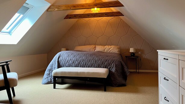 Un dormitorio con techo inclinado, una cama con cubrecama acolchado, un banco, un aparador y una ventana.
