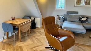 Una sala de estar acogedora con una mesa de comedor de madera, un sillón de cuero marrón, un sofá gris y una mesita auxiliar con una lámpara.