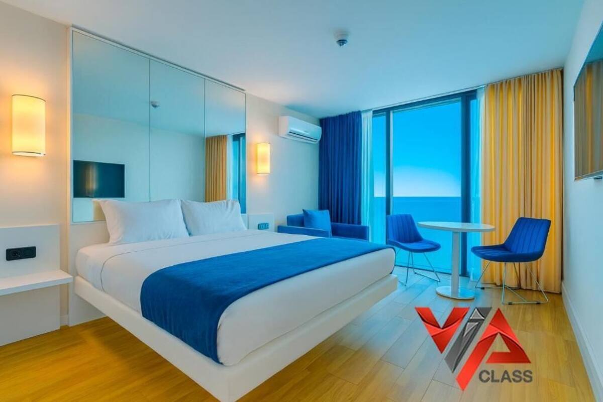 Una habitación de hotel moderna con una cama grande, una zona de estar con sillones, una pequeña mesa y un ventanal con cortinas.