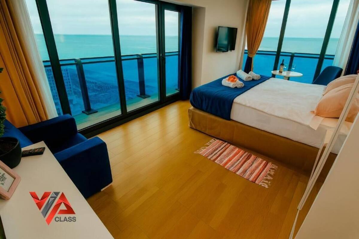 Habitación de hotel con un ventanal grande que ofrece vistas al mar, una cama con colcha azul, una mesita con una botella y vasos, y un sofá azul.