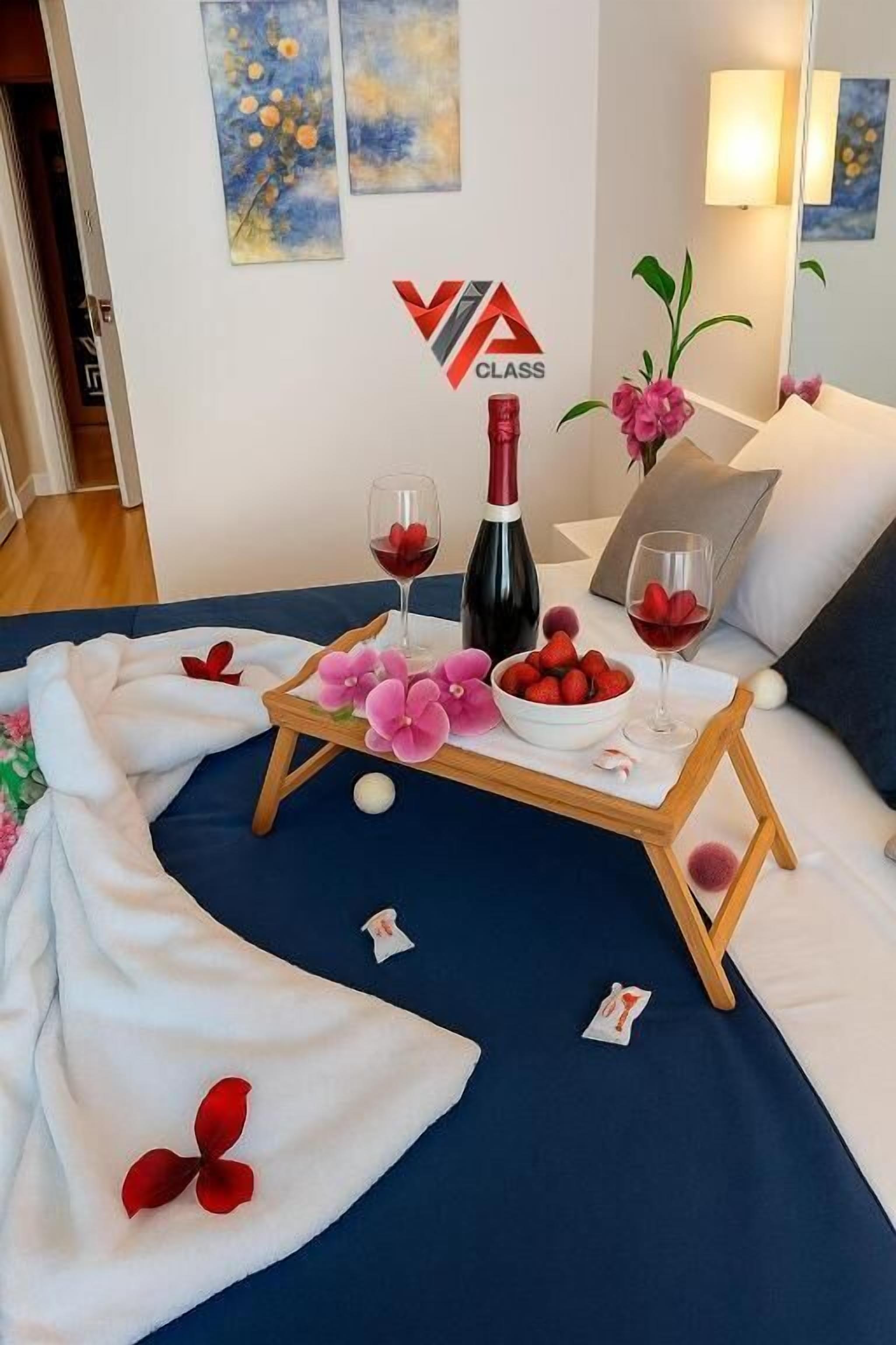 A bedroom with a bed, a bedside table with a wine bottle and glasses, a bowl of strawberries, and a floral arrangement.