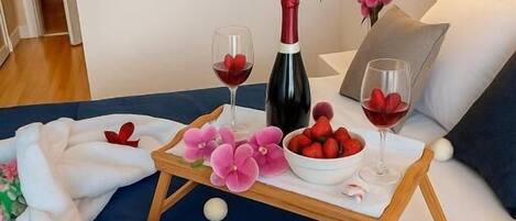 A bedroom with a bed, a bedside table with a wine bottle and glasses, a bowl of strawberries, and a floral arrangement.