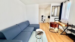 A modern living room with a blue sofa, a small round table, and a wooden coffee table. There is a kitchen area in the background with a dining table and chairs. - Superb Studio teletravail - Rooftop Paris (Saint-Ouen-sur-Seine)