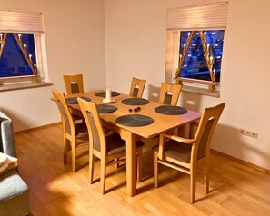 A dining area with a wooden table and chairs, a candle centerpiece, and windows with a view of the city at night. - Holiday Apartment De Gruusse (Mildenau)