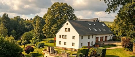 Ein großes, weißes Haus mit geneigtem Dach, umgeben von einem gepflegten Garten und Bäumen.