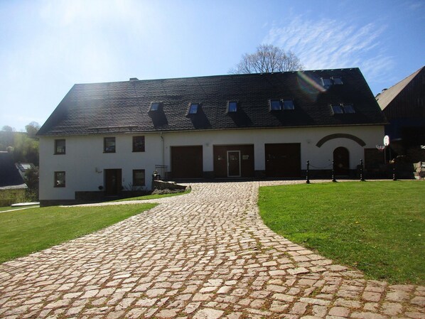 A building with a paved driveway leading to a front door, surrounded by a grassy lawn. - Holiday Apartment De Gruusse (Mildenau)
